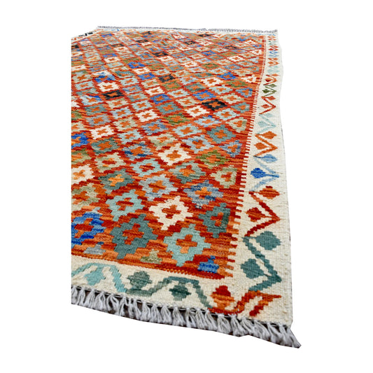 Angled view of the Chobi Kilim rug showing orange diamonds, teal details, an ivory patterned border and white fringe