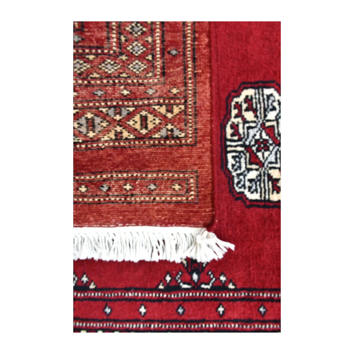 Folded end of Bokhara runner showing its patterned reverse beside the red front, part of an ivory gul and white fringe