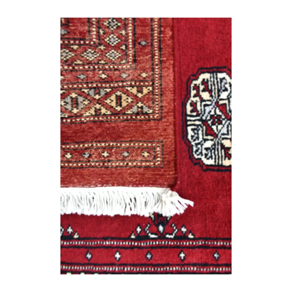 Folded end of Bokhara runner showing its patterned reverse beside the red front, part of an ivory gul and white fringe