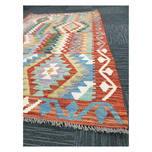 Angled view of Afghan Chobi Kilim rug showing terracotta border, fringe, and geometric flatweave detail, 180 x 122 cm