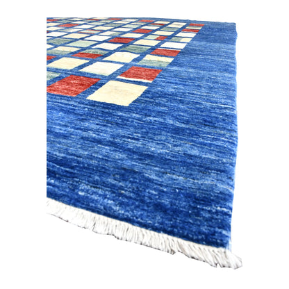 Angled detail of the Afghan Gabbeh rug showing its broad blue field, colour-block grid and white fringe