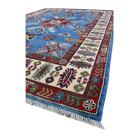 Angled view of Afghan Kazak wool rug showing its blue field, red medallion, ivory botanical border and pale fringe