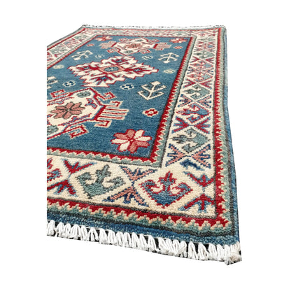 Angled corner and fringe view of Afghan Kazak wool rug showing blue field, cream border, red accents and light fringe