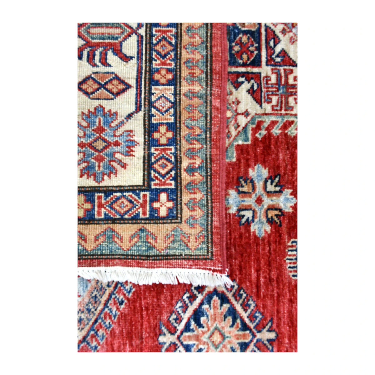 Folded corner of Afghan Super Kazak rug showing the detailed reverse weave, layered border and ivory fringe over the patterned face