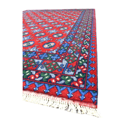 Angled view of Afghan Turkoman wool rug showing its red gul field, blue patterned borders and white fringe