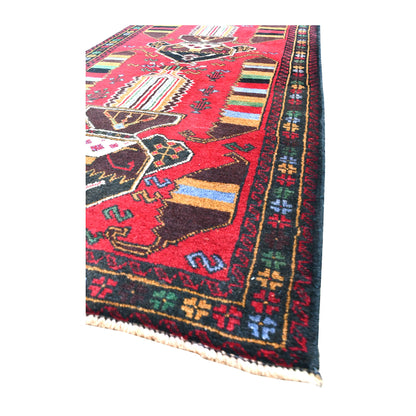 Angled view of Baluchi rug showing its red geometric field, dark patterned borders and short cream fringe
