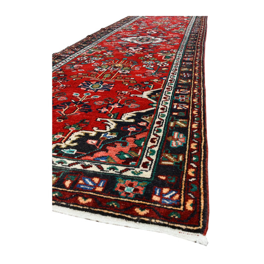 Angled corner of Persian Hamadan wool runner showing the red field, layered dark borders and short ivory fringe